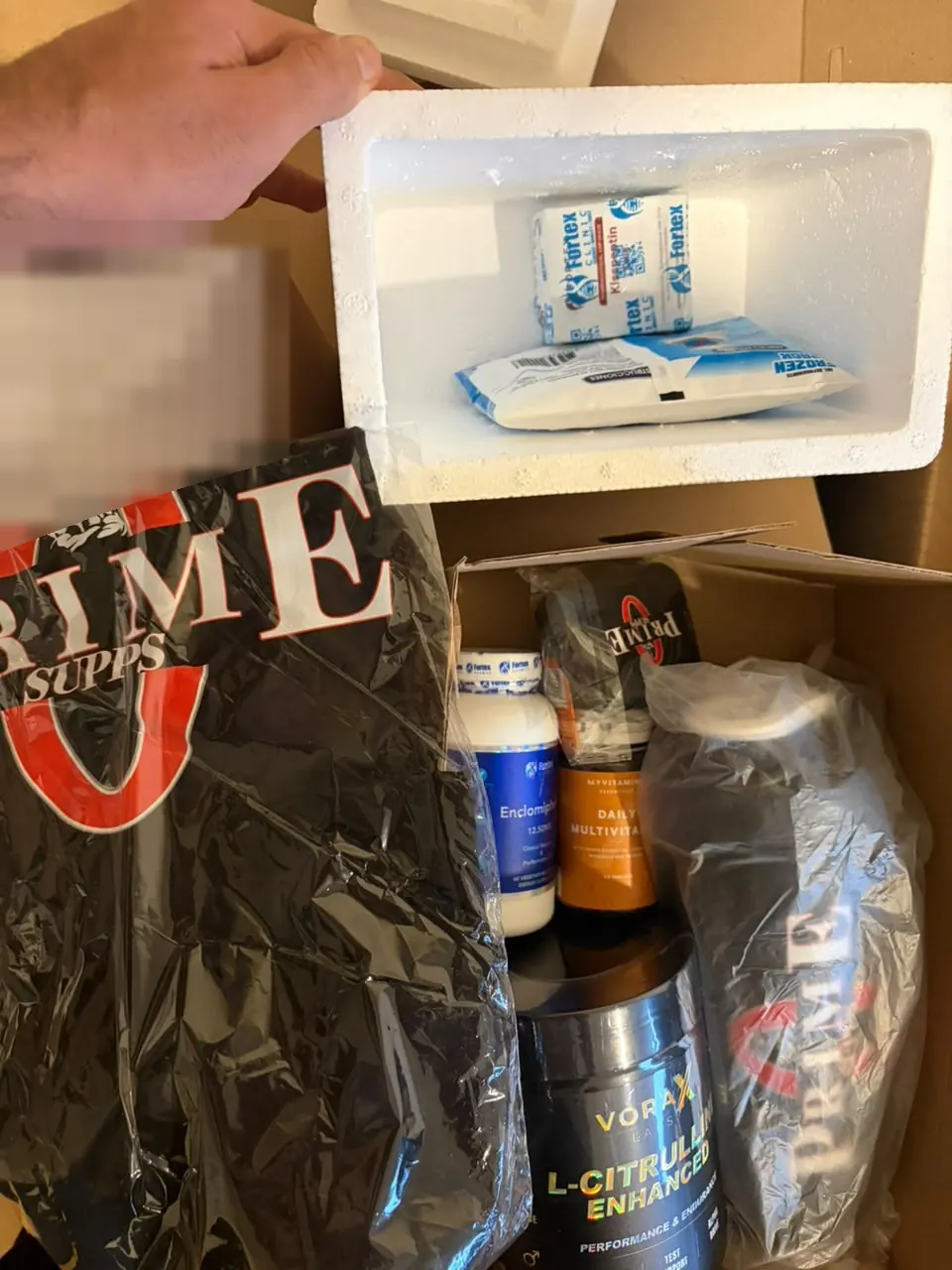 Caja de un pedido con péptidos en tecnopor y gel refrigerante, suplementos y polo Prime; la hoja de despacho está pixelada